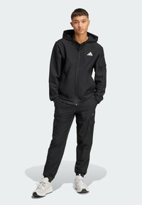 Black zip-up hoodie with a front logo, paired with cargo pants featuring side pockets. Worn with light-colored sneakers.