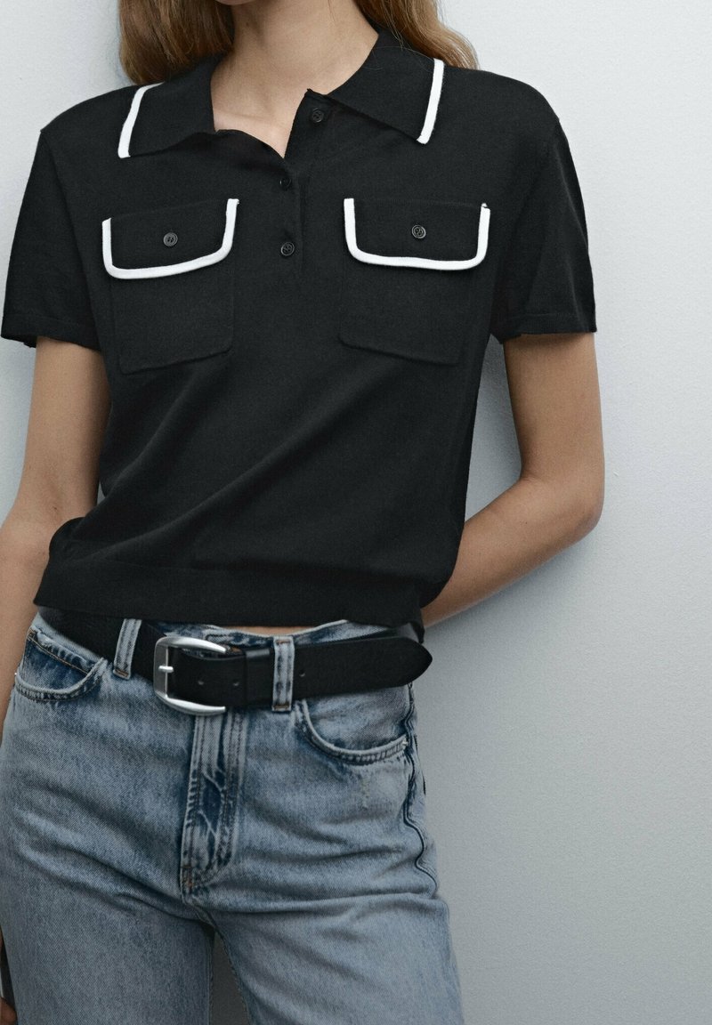 Femme portant un polo noir à manches courtes avec des poches bordées de blanc, rentré dans un jean bleu clair taille haute avec une ceinture noire.