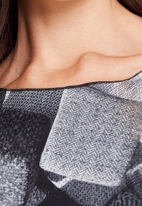 Czarno-biała tkanina w wzory z rybimi ośćmi i kształtami geometrycznymi, gładka w dotyku, z szerokim dekoltem, częściowo widoczna na ramieniu.