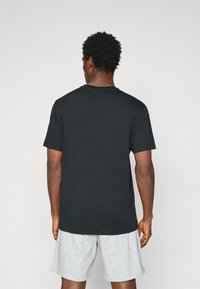 Schwarzes Baumwoll-T-Shirt mit Rundhalsausschnitt und kurzen Ärmeln, von hinten gesehen. Getragen mit grauen Shorts. Einfaches Design, keine sichtbaren Muster.