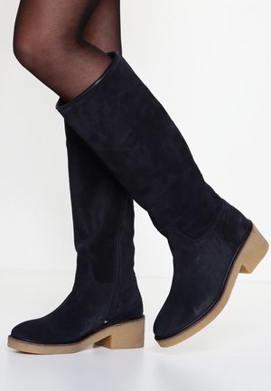 Nogi w czarnych zamszowych kozakach za kolano z jasnobrązowymi, kwadratowymi obcasami, stojące na białym tle.