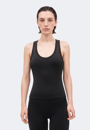 Ung person med kort vått hår har på seg en tettsittende svart singlet og svarte bukser, står rett opp mot en ensfarget lys bakgrunn.
