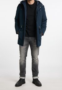 Uomo che indossa un parka con cappuccio blu navy, una t-shirt nera, jeans grigi sbiaditi con risvolti e scarpe nere, in piedi con le mani in tasca su uno sfondo bianco.