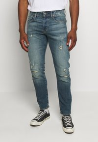 Man i slitna blå jeans, vita strumpor och svarta låga sneakers står mot en enkel ljus bakgrund.