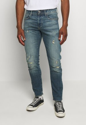 Hombre con jeans azules desgastados, calcetines blancos y zapatillas negras de caña baja, de pie contra un fondo claro y liso.