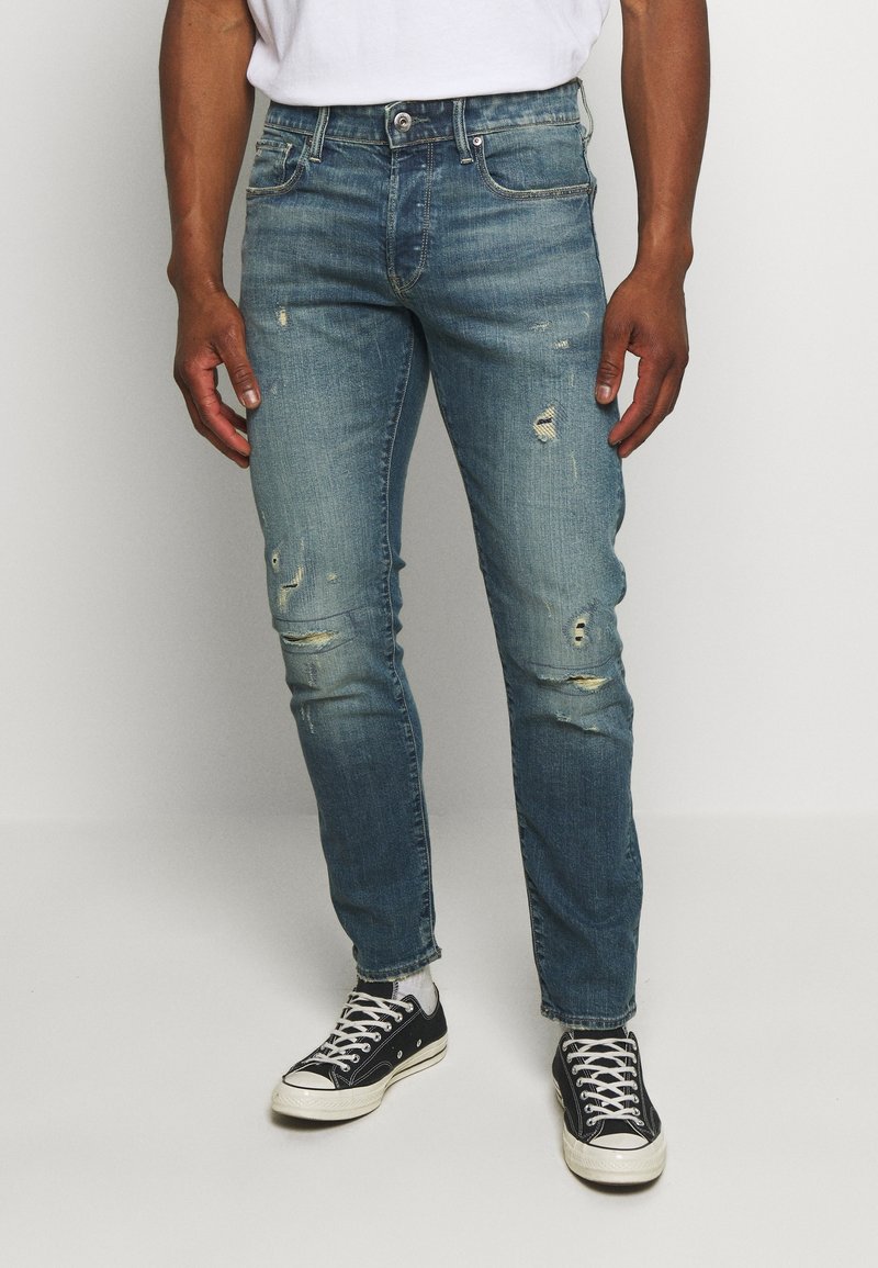 Man i slitna blå jeans, vita strumpor och svarta låga sneakers står mot en enkel ljus bakgrund.
