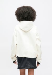 Λευκό υπερμεγέθες μπουφάν με κουκούλα και ελαστικά μανίκια, εμφανιζόμενο από πίσω. Συνδυασμένο με μαύρη φούστα για ένα στιλ με στρώσεις. Λείο ύφασμα.