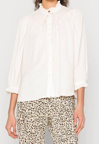 Blusa blanca abotonada con cuello y puños con volantes, con cierre de lazo. Combinada con pantalones beige plisados con manchas negras.