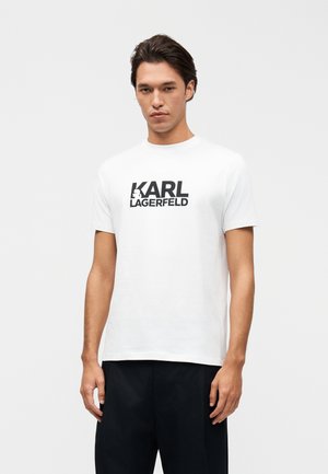 Moški model stoji, oblečen v belo kratkoročno majico s črnim napisom "KARL LAGERFELD" in črne hlače na enobarvnem ozadju.