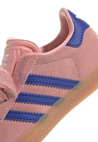 Gros plan sur une basket Adidas en daim rose avec des rayures bleues, une bride à scratch rose, un talon bleu et une semelle en gomme translucide.