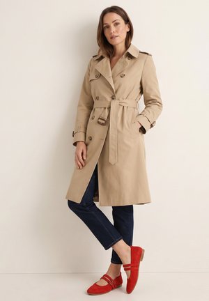 Frau trägt einen beigen Trenchcoat mit Gürtel, dunkelblaue Jeans und rote Mary Jane Ballerinas, steht vor einer einfachen hellen Wand.
