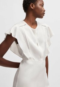 Biała sukienka z krótkimi falbaniastymi rękawami, subtelnym detalem w kształcie klucza w dekolcie, dopasowanym krojem oraz gładką, miękką teksturą materiału.