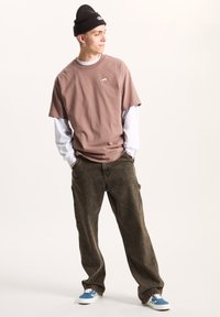 T-shirt marrone a collo giro stratificata su una maglietta bianca a maniche lunghe, abbinata a pantaloni cargo scuri e sneakers blu con motivo. Indossa un berretto nero.