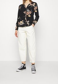 Langärmliges schwarzes Blumenhemd mit V-Ausschnitt, kombiniert mit weißen, locker sitzenden Hosen und schwarzen High-Top-Sneakern.