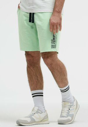 Mann trägt hellgrüne Shorts mit schwarzen Kordeln und Schriftzug, weiße Socken mit schwarzen Streifen und weiße Sneakers, steht auf einfachem Hintergrund.