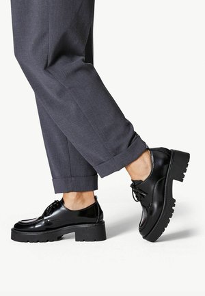 Chaussures vernies noires avec un bout rond, semelle épaisse et chunky, et un design à lacets, associées à un pantalon gris à revers.