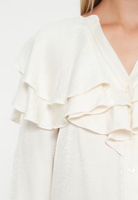 Crème blouse met gelaagde ruffles aan de voorkant, knoopsluiting en een gladde, lichtjes gestructureerde stof.