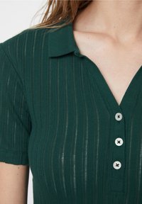 Zbliżenie na osobę w ciemnozielonej, prążkowanej koszulce z krótkim rękawem, z kołnierzykiem i trzema białymi guzikami z przodu.