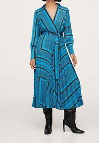 Vestido envolvente azul con rayas geométricas negras, mangas largas, cuello abotonado y cinturón en la cintura. La tela tiene una textura sutil. Botas negras hasta la rodilla.