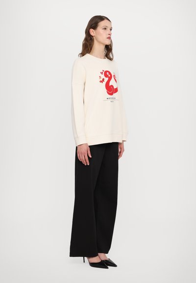 Cremfarbener Pullover mit einem roten grafischen Design, das eine Schlange und Schmetterlinge zeigt, kombiniert mit schwarzen weitgeschnittenen Hosen und spitzen schwarzen Absatzschuhen.