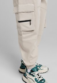 Béžové cargo kalhoty s kapsami na zip a chlopněmi, doplněné o bílé a tyrkysové tenisky na šedém pozadí.