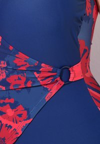 Maillot de bain au design ajusté mettant en valeur une base bleu marine et un motif floral rouge sur un côté, maintenu par un accent métallique circulaire.