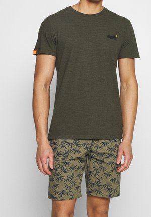 Mand iført olivengrøn kortærmet T-shirt med lille logo på brystet og olivengrønne shorts med mørkt bladmønster.