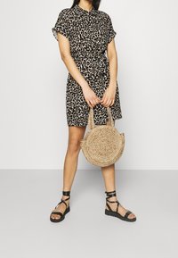 Sukienka koszulowa w panterkę z krótkim rękawem i paskiem w talii, zestawiona z czarnymi sandałami na ramiączkach oraz okrągłą, plecioną torebką.