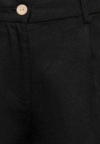 Pantalon noir avec poche avant et un seul bouton clair à la taille, montrant la texture du tissu et les détails des coutures.