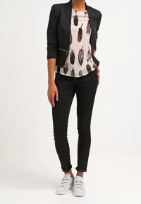 Blazer nero su misura sopra una blusa chiara con stampa di piume, abbinato a jeans skinny neri e sneaker grigie. Texture lisce ovunque.