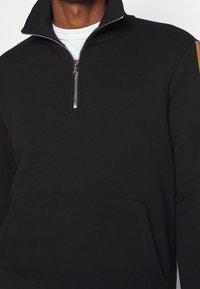Pullover nera a zip corta realizzata in tessuto soffice. Presenta una tasca a marsupio frontale, una zip argentata e un accento di colore con strisce sulle maniche.
