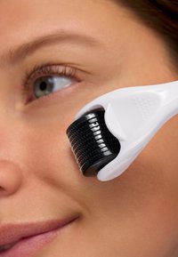 Svart derma roller med fina nålar fästa vid ett vitt handtag, används på huden nära kinden och visar dess rundade form och textur.