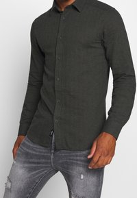 Camicia a maniche lunghe verde scuro in tessuto texturizzato, con colletto alla francese e chiusura frontale con bottoni. Indossata con jeans grigi strappati.