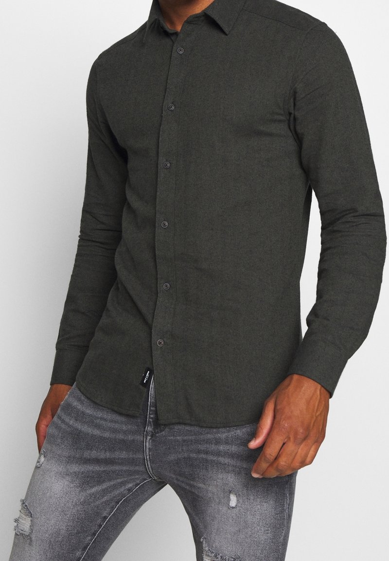 Camicia a maniche lunghe verde scuro in tessuto texturizzato, con colletto alla francese e chiusura frontale con bottoni. Indossata con jeans grigi strappati.