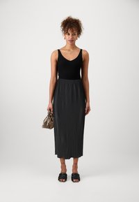 Top scurt din tricot negru, asociat cu o fustă midi plisată de culoare gri închis. Sandale negre cu talpă joasă și o geantă mică, de culoare taupe, cu detalii plisate.