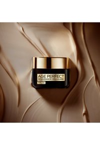 Pot noir avec un couvercle doré, étiqueté "AGE PERFECT RENAISSANCE CELLULAIRE FPS 30." Fond avec une texture de crème beige lisse.