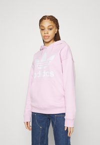 Lys rosa hettegenser laget av bomull, med en stor hvit Adidas-logo og en kengurulomme foran. Mansjetter på ermene og avslappet passform.