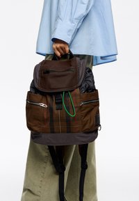 Rjava karirana nahrbtnik s temnim zgornjim pokrovom, žepki na zadrgo, zelenimi detajli in prilagodljivimi naramnicami. Narejen iz trpežnih materialov.