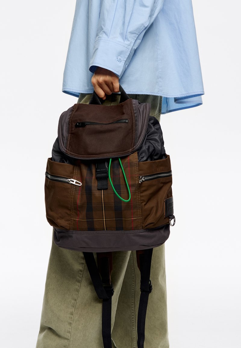 Rjava karirana nahrbtnik s temnim zgornjim pokrovom, žepki na zadrgo, zelenimi detajli in prilagodljivimi naramnicami. Narejen iz trpežnih materialov.