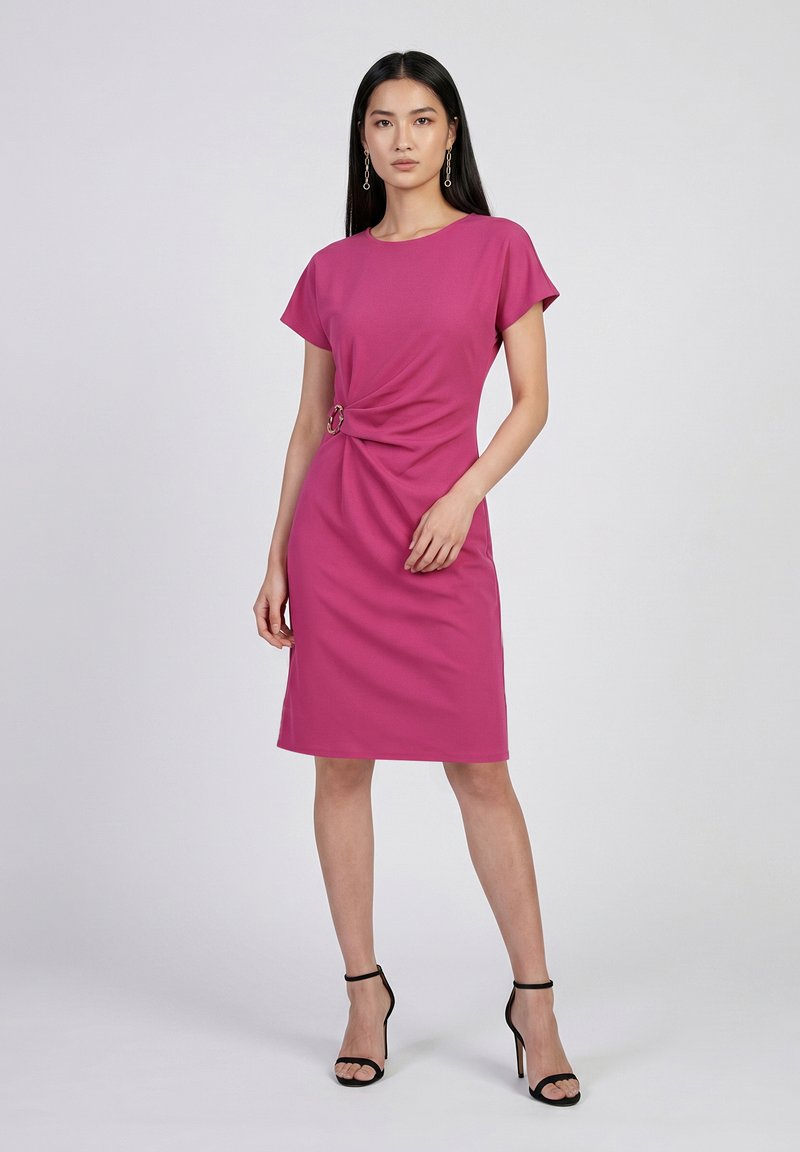 Femme aux longs cheveux noirs portant une robe rose mi-longue avec un détail de boucle latérale et des sandales noires à talons hauts, debout devant un fond uni.