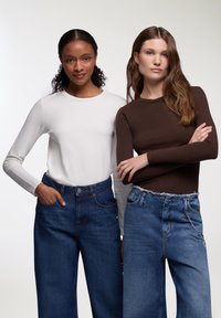Due modelle indossano magliette a maniche lunghe, una bianca e l'altra marrone, abbinate a jeans in denim blu a vita alta.