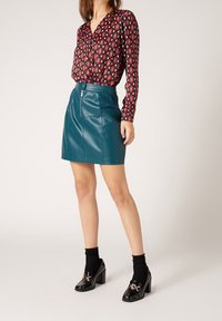 Rot-schwarz gemusterte Bluse mit langen Ärmeln, kombiniert mit einem taubenblauen Leder-Minirock und schwarzen Lackleder-Pumps mit goldenen Akzenten.