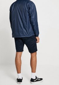 Chaqueta ligera navy con una textura suave, combinada con pantalones cortos navy. Zapatillas negras con suelas blancas y calcetines completan el atuendo.