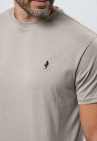 Približna snimka muškarca koji nosi svijetlo bež majicu kratkih rukava s malom crnom izvezenom siluetom osobe u baloneru na prsima.