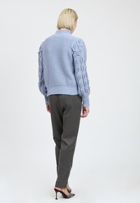 Helles blaues Strickpullover mit strukturierten Kabelstrick-Ärmeln, hohem Kragen und geripptem Saum, kombiniert mit grauen Hosen und gemusterten Absätzen.