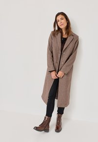 Cappotto di lana marrone con una silhouette lunga, due bottoni e colletto a scialle, abbinato a jeans neri e stivali stringati marroni.