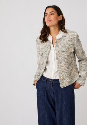 Mujer con chaqueta beige texturizada con botones, blusa blanca con cuello con volantes y vaqueros oscuros de talle alto y pierna ancha, manos en los bolsillos.