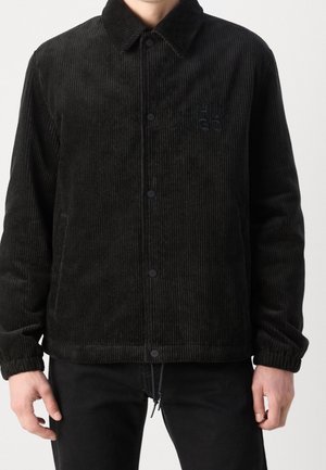 Schwarze Cordjacke mit Druckknöpfen und elastischen Bündchen, getragen von einer Person, die vor einem einfarbigen Hintergrund steht.