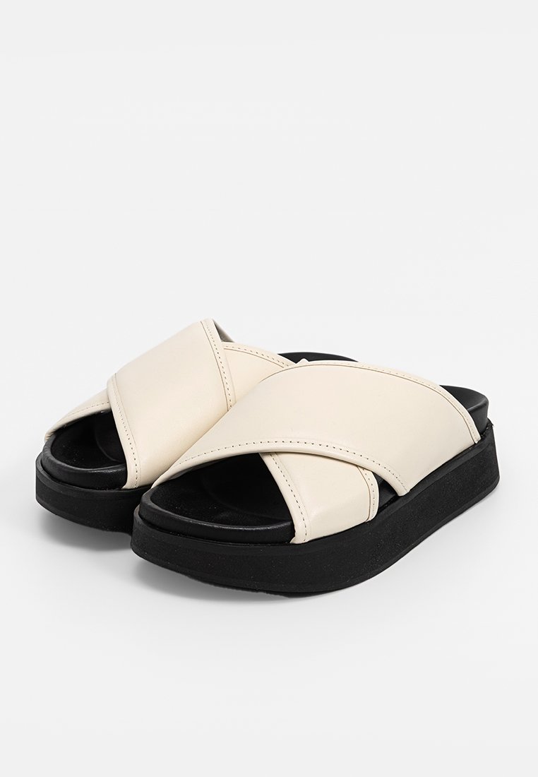 Sandales à brides croisées crème avec une semelle noire texturée, présentant un dessus en cuir lisse, un design à bout ouvert et des détails de couture subtils.