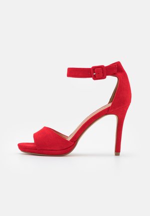 Sandale à talon haut en suédine rouge avec bout ouvert, bride de cheville et boucle sur fond blanc.